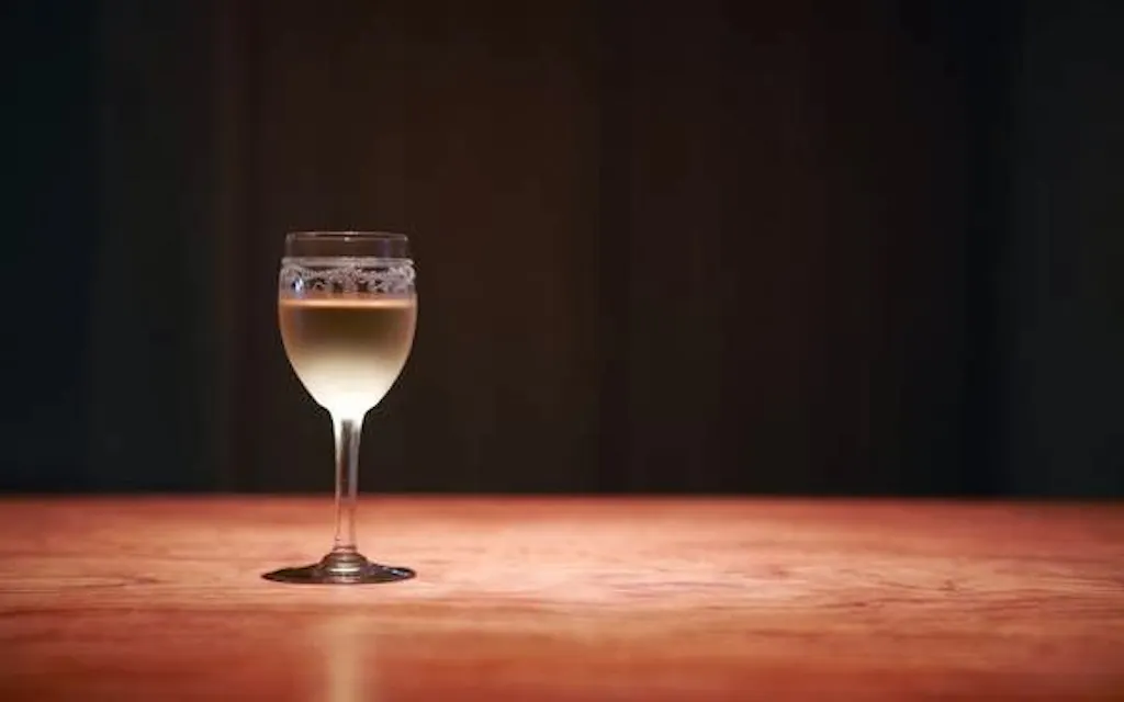 Sayura Vins Fins A single glass of white wine sits on a wooden table against a dark, blurred background. The wine appears to be chilled, with condensation forming on the upper part of the glass. The lighting highlights the golden hue of the wine.