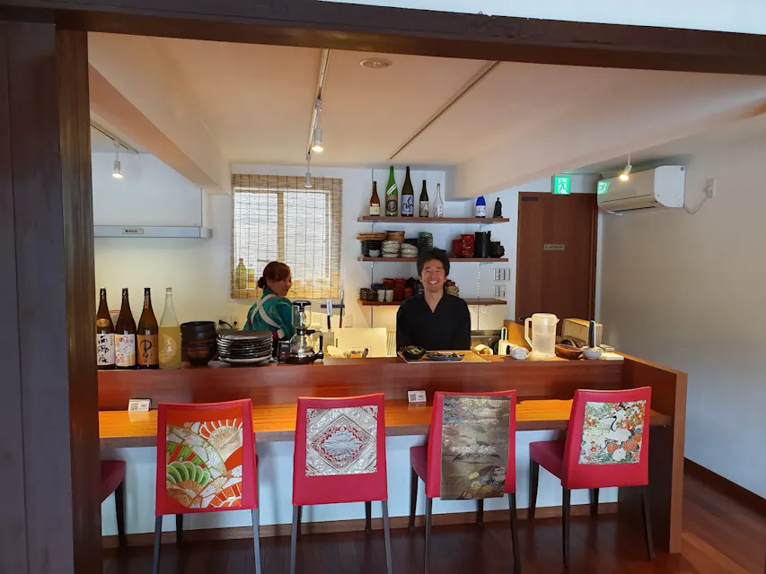 Shishin Samurai Cafe & Bar A cozy Japanese restaurant features a bar with four colorful, upholstered stools. Behind the bar, a smiling man and a woman prepare drinks and food. Shelves with plates, glasses, and bottles of sake are visible in the background. Warm lighting enhances the ambiance.