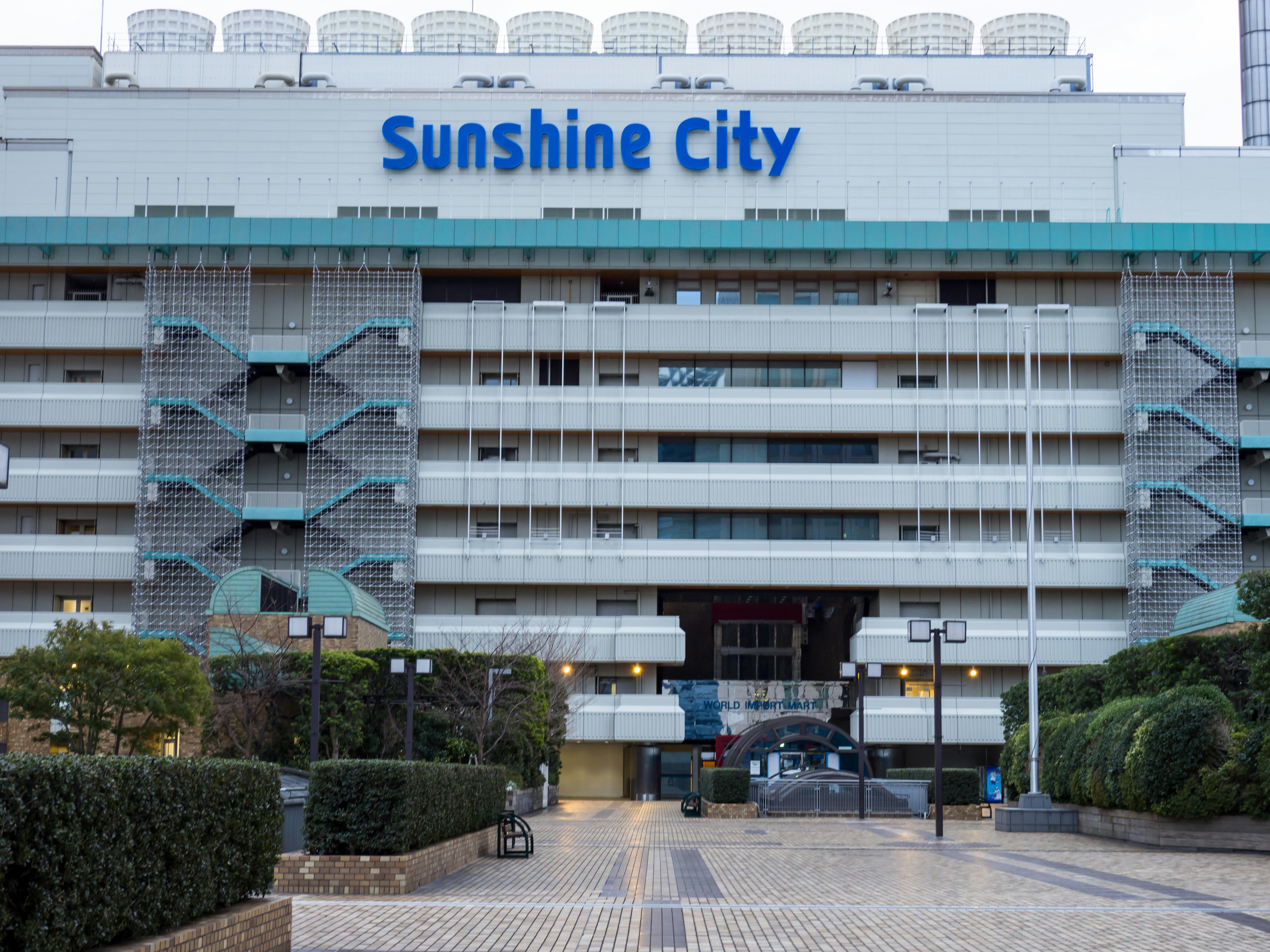 A large building with the sign "Sunshine City" on the upper facade, featuring multiple levels, patterned staircases, and an open plaza area with greenery and brick pavement in front.