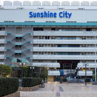 Sunshine City Ikebukuro A large building with the sign "Sunshine City" on the upper facade, featuring multiple levels, patterned staircases, and an open plaza area with greenery and brick pavement in front.