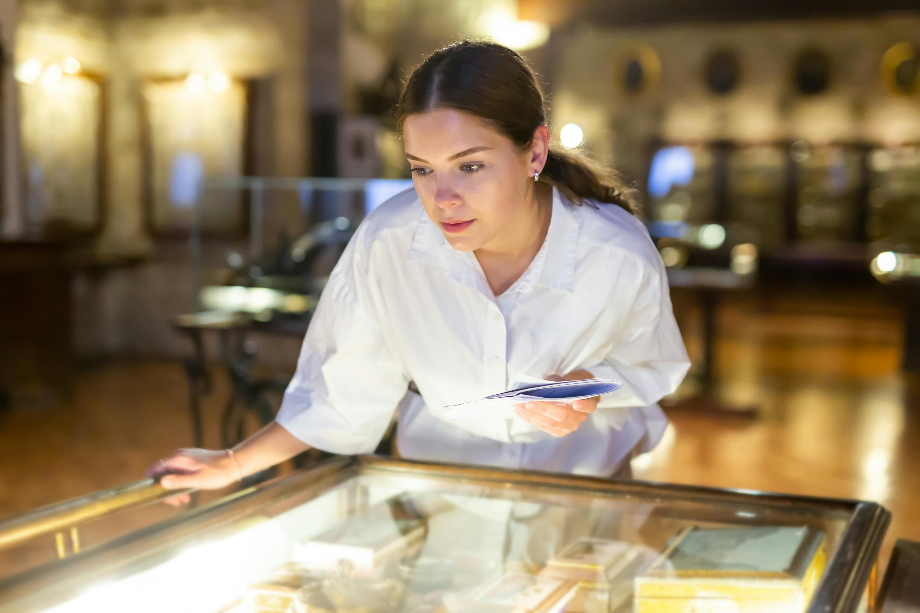 A woman in a white shirt closely examines items displayed in a glass case at a museum, holding a brochure or pamphlet in one hand. The setting is softly lit with blurred exhibits in the background.