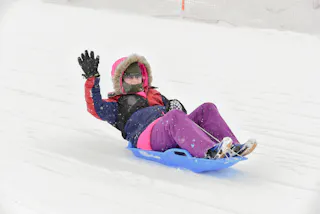 A person bundled in winter clothes, including a hooded jacket, gloves, and sunglasses, sleds down a snowy hill on a blue sled, waving cheerfully with one hand. Snow is falling lightly around them.