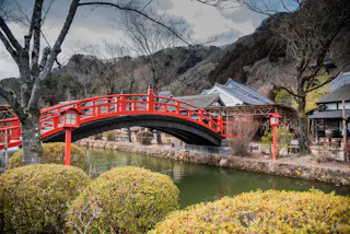 A red arched bridge crosses a narrow river in a peaceful Japanese garden, surrounded by green bushes, leafless trees, traditional buildings, and distant wooded hills under a cloudy sky.