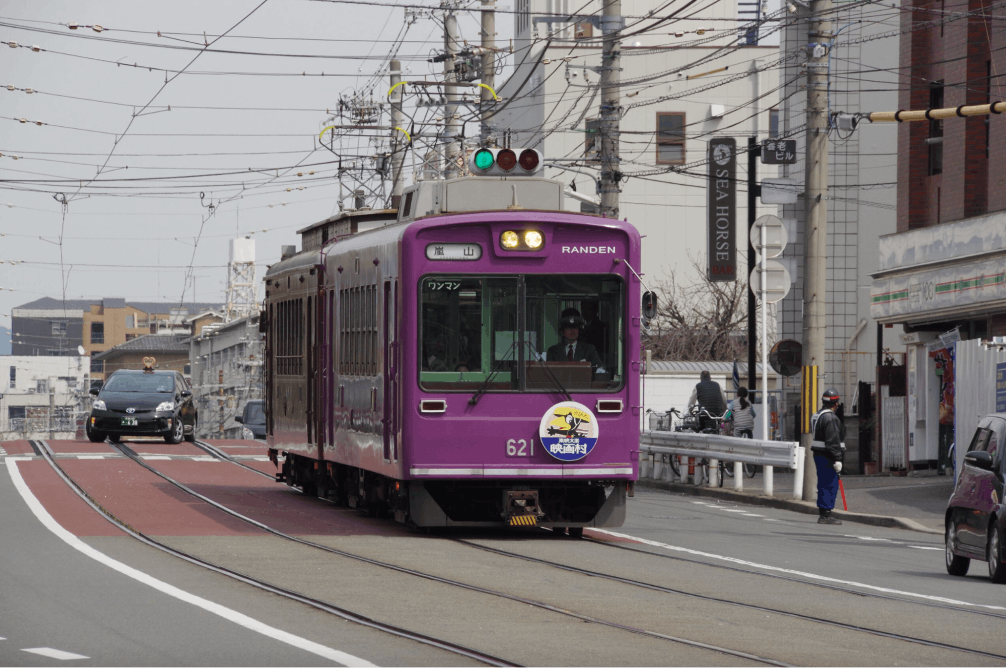 A purple streetcar with the number 621 travels along tracks on a city street, passing buildings, cars, and overhead wires. The tram displays a decorative sign and a green traffic signal is visible above.