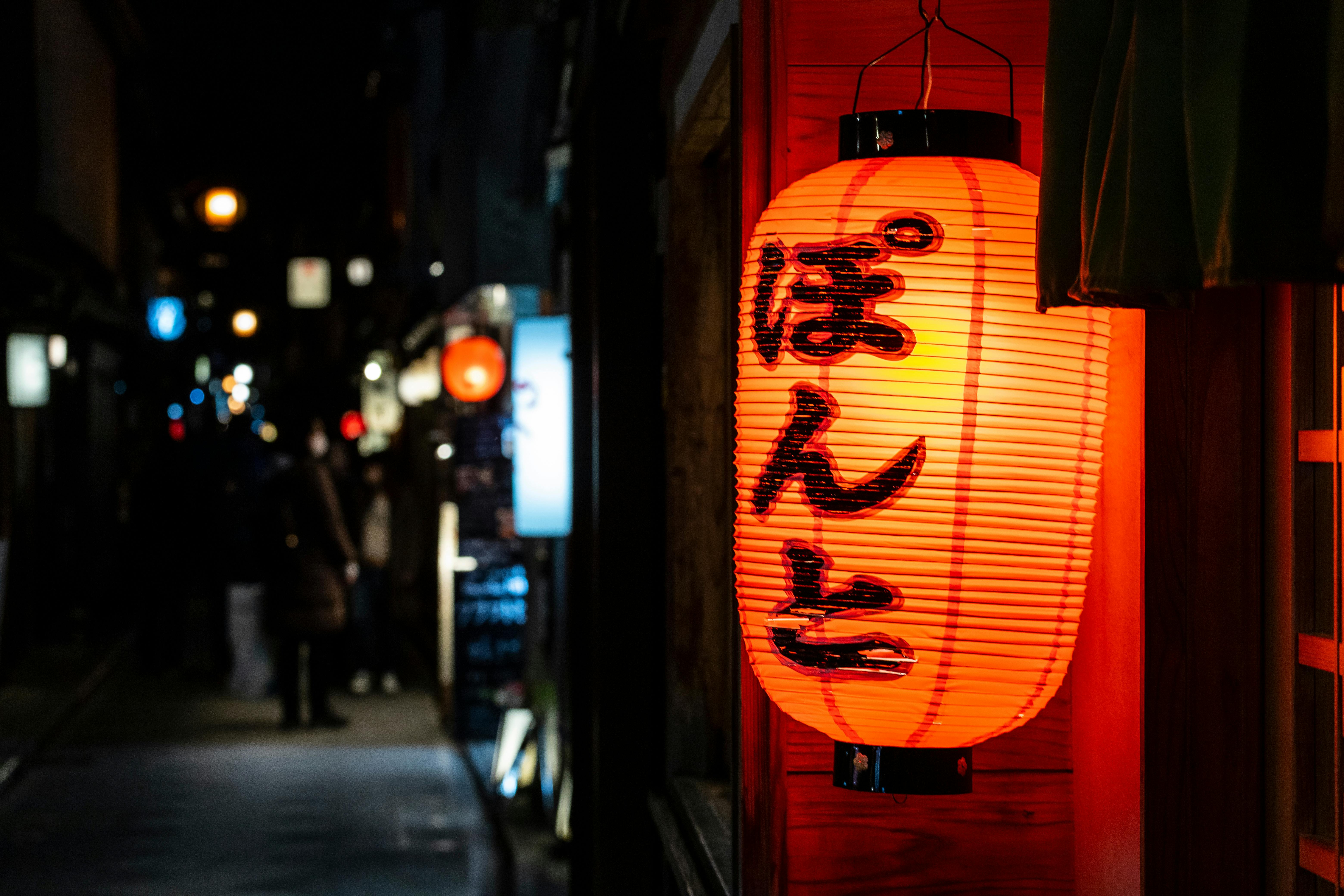 A glowing red Japanese paper lantern with black kanji characters hangs outside a building at night, illuminating a narrow street with more lanterns and blurred pedestrians in the background.