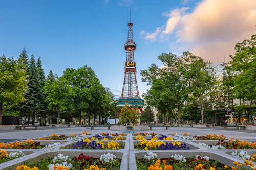 The Sapporo TV Tower rises behind colorful flower beds and green trees in a city park, with a blue sky and some clouds in the background.