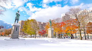 Two statues stand on pedestals in a snowy park with colorful autumn trees. Tall buildings are visible in the background under a partly cloudy blue sky.