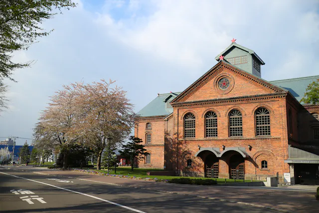 Red brick building with arched windows and star decorations, bordered by a quiet street and trees, under a partly cloudy sky.