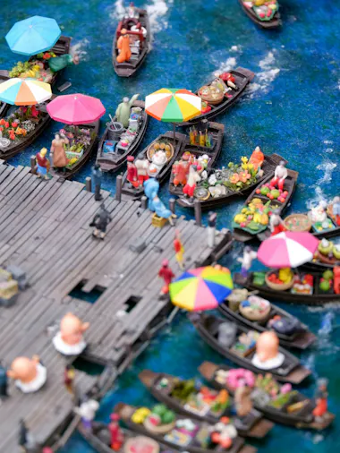 Aerial view of a busy floating market with colorful umbrellas, vendors selling goods from boats, and people walking on a wooden dock by vibrant blue water.