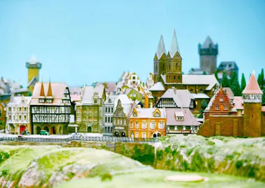 A detailed miniature model of a European town features colorful historic buildings, church towers, and a castle, set against a bright blue sky with green grassy foreground.