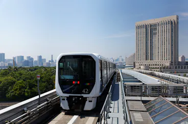 A modern white train travels on an elevated track in an urban area with a large building and city skyline in the background under a clear blue sky.
