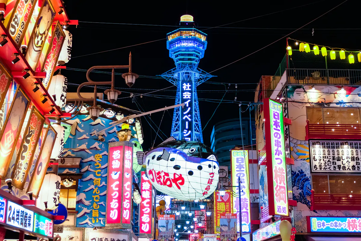 Night view of a vibrant street in Osaka, Japan, with colorful neon signs and billboards lining the buildings. In the background, Tsutenkaku Tower is illuminated in blue, towering above the lively shopping district.