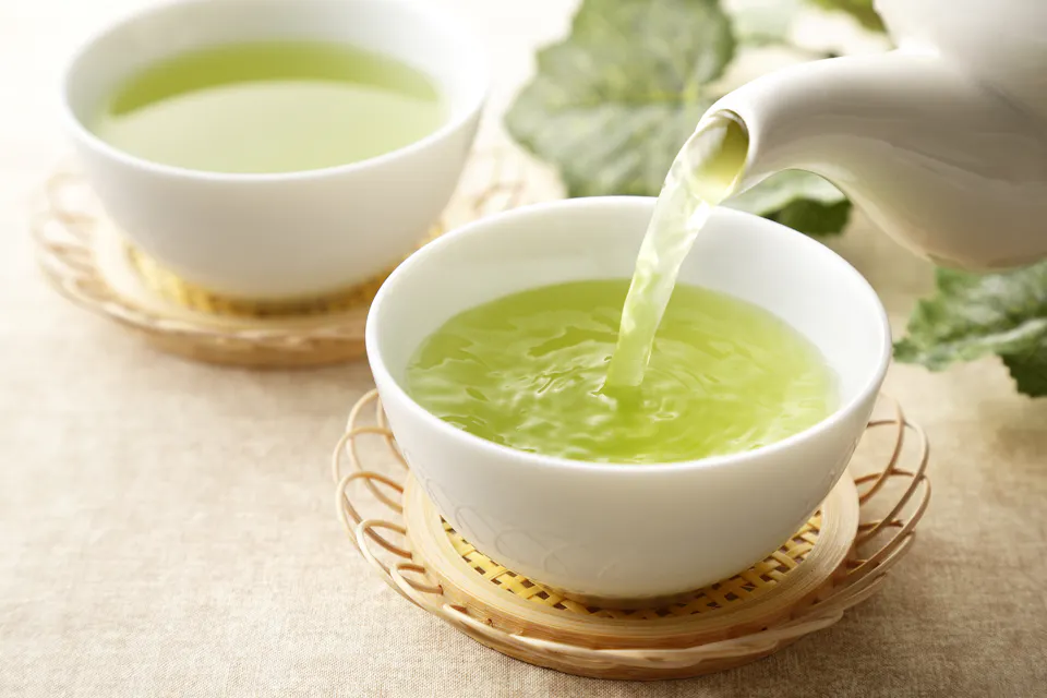 A teapot pours green tea into a white cup, placed on a woven coaster. Another cup of green tea sits nearby. The background features soft-focus green leaves, creating a calming, natural atmosphere.