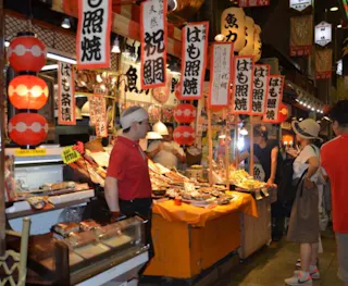 A bustling indoor Japanese market with food stalls, red lanterns, and signs in Japanese. Vendors and shoppers interact near displays of fresh seafood and local delicacies.