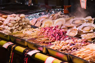 Trays of various skewered seafood, including octopus, scallops, and shellfish, are displayed at a market stall under bright lighting, with prices visible on small signs.