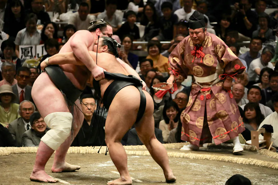 Two sumo wrestlers grapple in a ring during a match, surrounded by an audience seated closely. A referee in traditional attire watches closely, poised with a fan. The wrestlers wear traditional sumo mawashi belts, and one has a bandaged knee.