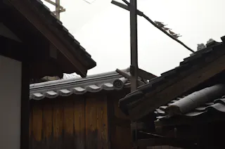 Traditional wooden buildings with dark tiled roofs, featuring a bamboo pole structure above. The scene appears calm and misty, highlighting the textures and details of the wooden walls and roof tiles.