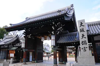 A traditional Japanese temple entrance featuring a large wooden gate with ornate carvings and lanterns. There is a tall stone pillar with Japanese characters inscribed on it. The architecture is characterized by curved, tiled roofs. Trees and buildings are visible in the background.