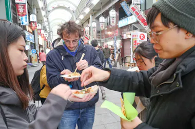 Four people stand in a covered outdoor market, eating street food from trays with chopsticks. Colorful signs and shopfronts line the background. The atmosphere appears casual and social.