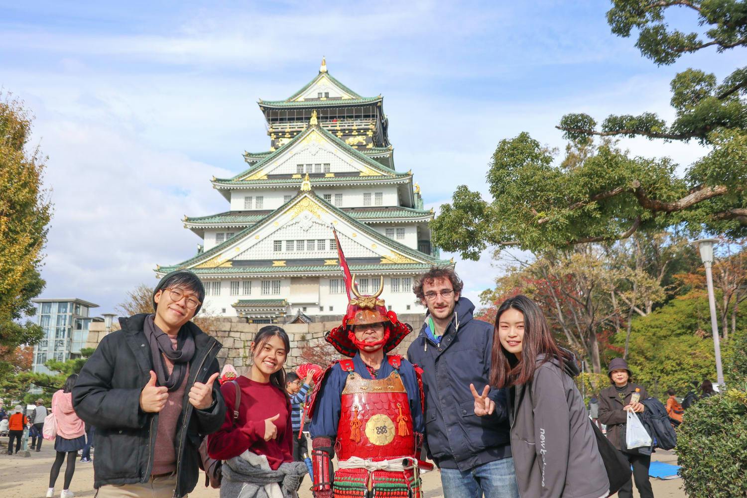 A group of four people poses with a person dressed as a samurai in red armor in front of Osaka Castle, surrounded by trees and other visitors on a clear day.