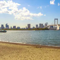 Odaiba Seaside Park A sandy beach borders calm water with a boat, city skyline, and a large suspension bridge in the background under a partly cloudy sky.