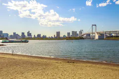 A sandy beach borders calm water with a boat, city skyline, and a large suspension bridge in the background under a partly cloudy sky.