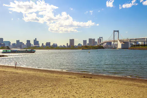 A sandy beach borders calm water with a boat, city skyline, and a large suspension bridge in the background under a partly cloudy sky.