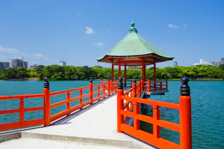 A red wooden pavilion with a green roof sits over blue water, connected by a walkway. Green trees and city buildings are visible in the background under a clear blue sky.