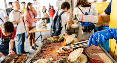 A person wearing blue gloves uses a blowtorch to cook shellfish on a grill, while a group of people stand and watch in a busy indoor market or food event.