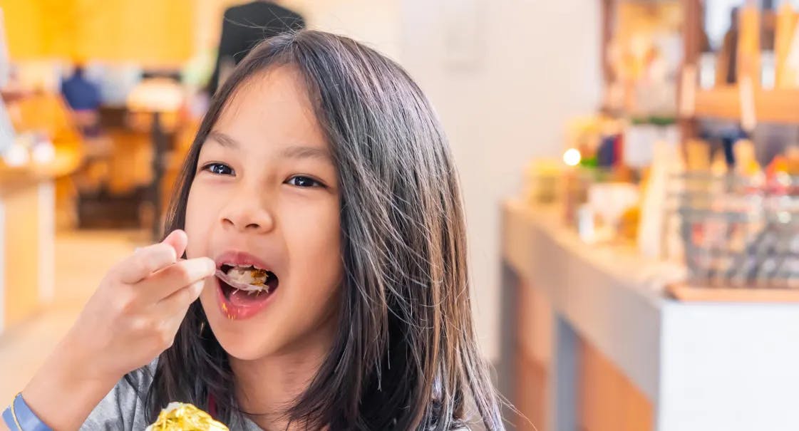 A young girl with long dark hair is eating food with a spoon, looking happy. She is indoors, with a blurred background of what appears to be a kitchen or dining area.