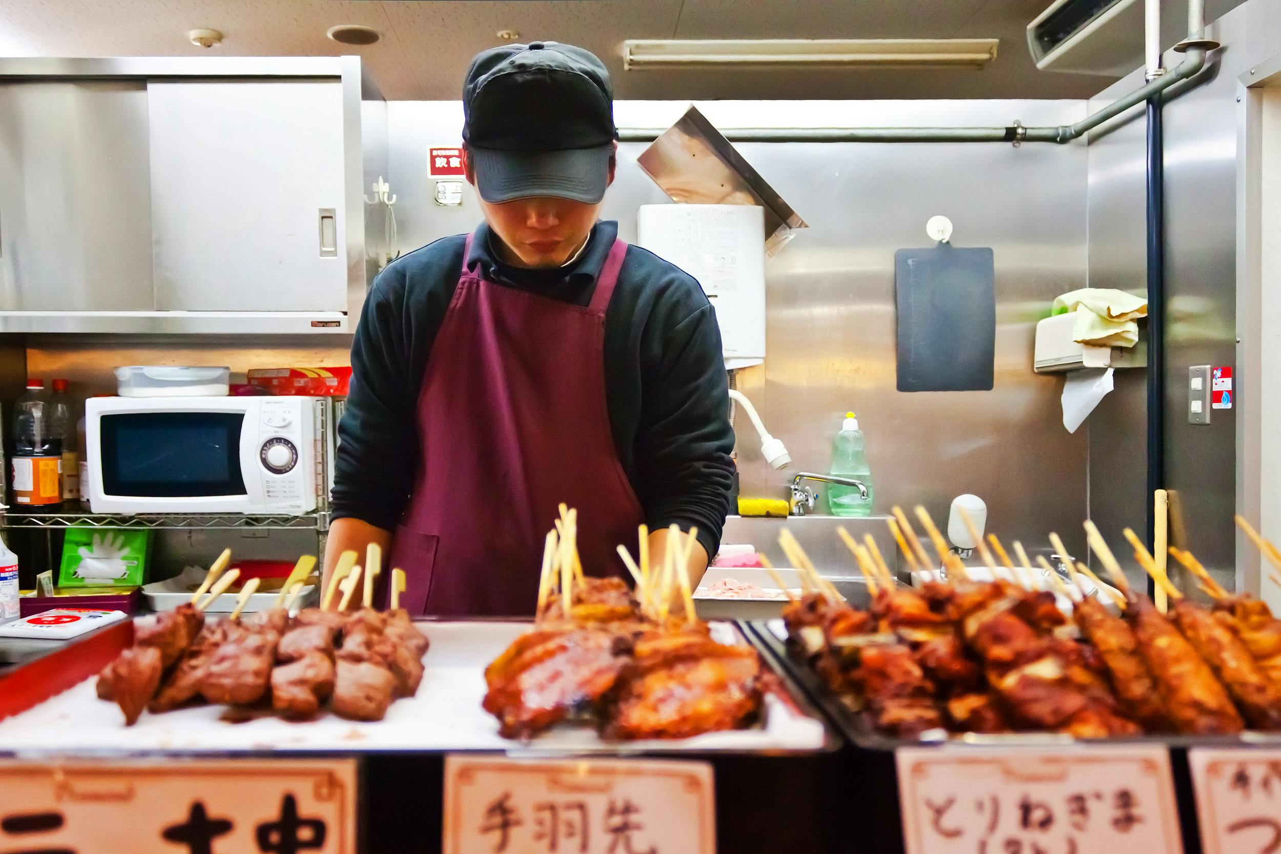 A vendor in a maroon apron and black cap arranges skewered grilled meats at a food stall, with trays of yakitori and handwritten signs, inside a kitchen with a microwave and utensils in the background.