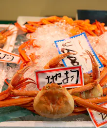 Omicho Market A display of fresh crabs on ice at a seafood market, with handwritten Japanese price tags placed on top of the crabs. The crabs have orange shells and long legs.