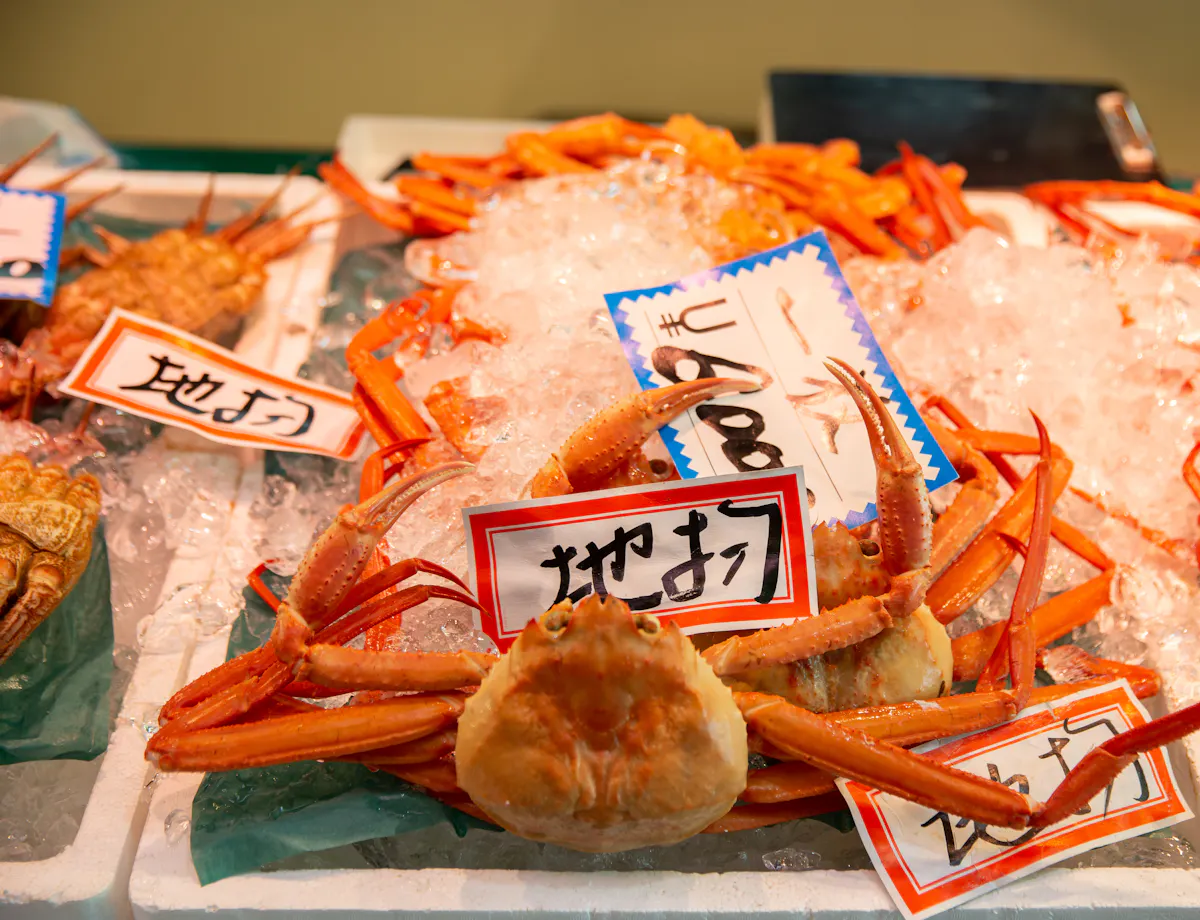 Omicho Market A display of fresh crabs on ice at a seafood market, with handwritten Japanese price tags placed on top of the crabs. The crabs have orange shells and long legs.