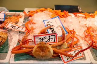 A display of fresh crabs on ice at a seafood market, with handwritten Japanese price tags placed on top of the crabs. The crabs have orange shells and long legs.