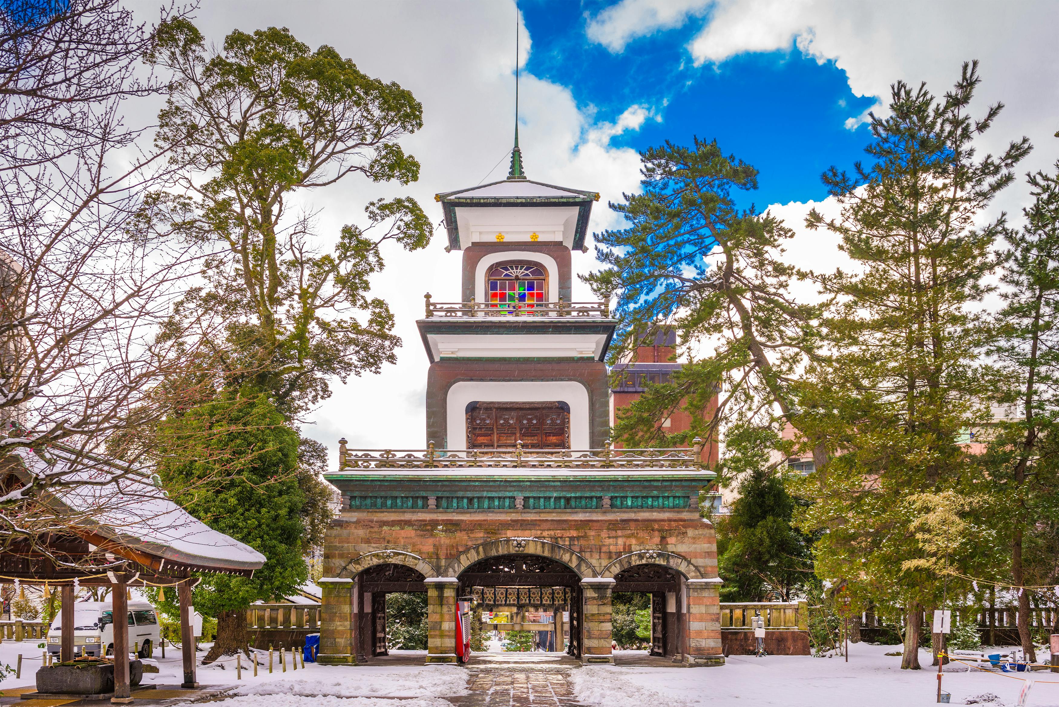A traditional Japanese temple gate with arched entrances stands amid snow-covered ground and tall trees, under a partly cloudy sky. The structure has intricate details and a central tower.