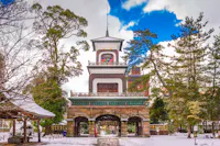 A traditional Japanese temple gate with arched entrances stands amid snow-covered ground and tall trees, under a partly cloudy sky. The structure has intricate details and a central tower.