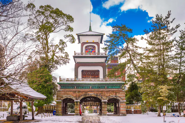 A traditional Japanese temple gate with arched entrances stands amid snow-covered ground and tall trees, under a partly cloudy sky. The structure has intricate details and a central tower.