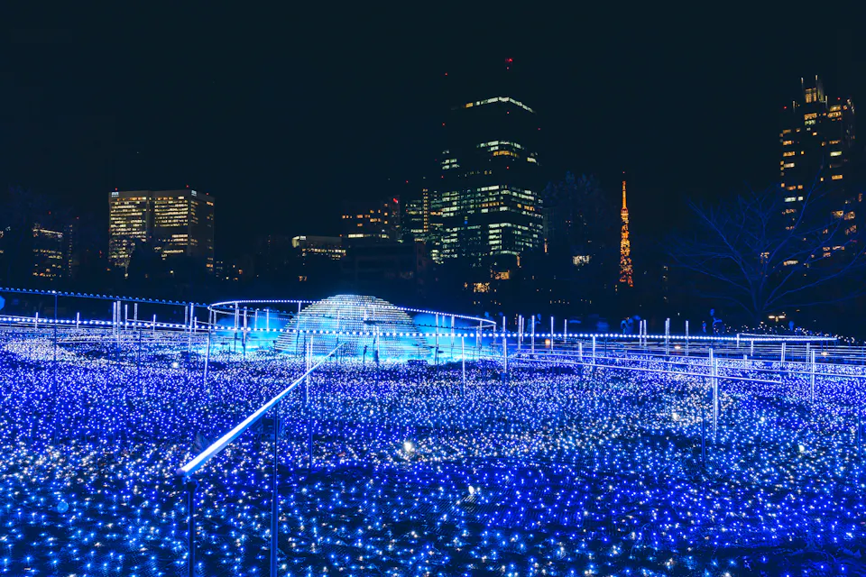 A cityscape at night featuring a dazzling display of blue LED lights on the ground, creating a sea-like effect. Skyscrapers and a tower with orange lights rise in the background, adding to the urban atmosphere.
