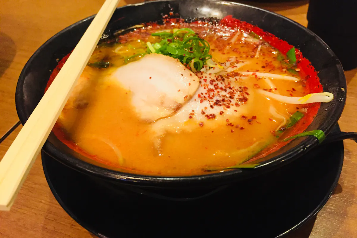Ramen in Osaka A bowl of ramen with a rich, orange-hued broth sits on a black plate. It is topped with slices of meat, green onions, bean sprouts, and chili flakes. A pair of wooden chopsticks rests across the bowl. The wooden table surface is visible in the background.
