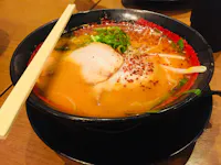 A bowl of ramen with a rich, orange-hued broth sits on a black plate. It is topped with slices of meat, green onions, bean sprouts, and chili flakes. A pair of wooden chopsticks rests across the bowl. The wooden table surface is visible in the background.