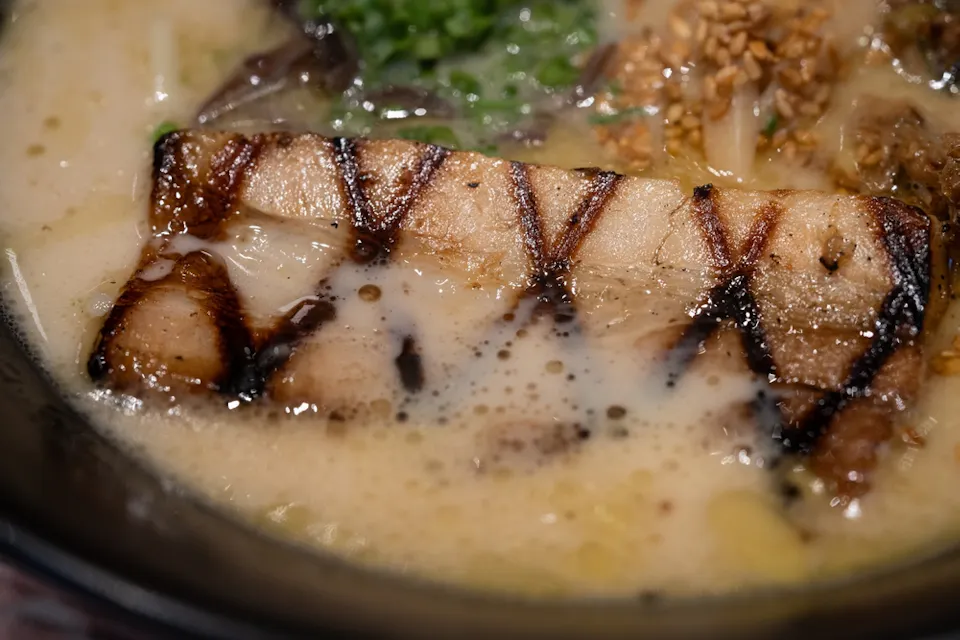 Close-up of a bowl of ramen with a thick, grilled pork belly slice as the centerpiece. The creamy broth shows bubbles and oil droplets with additional toppings like chopped green onions, mushrooms, and minced garlic adding texture and color to the dish.