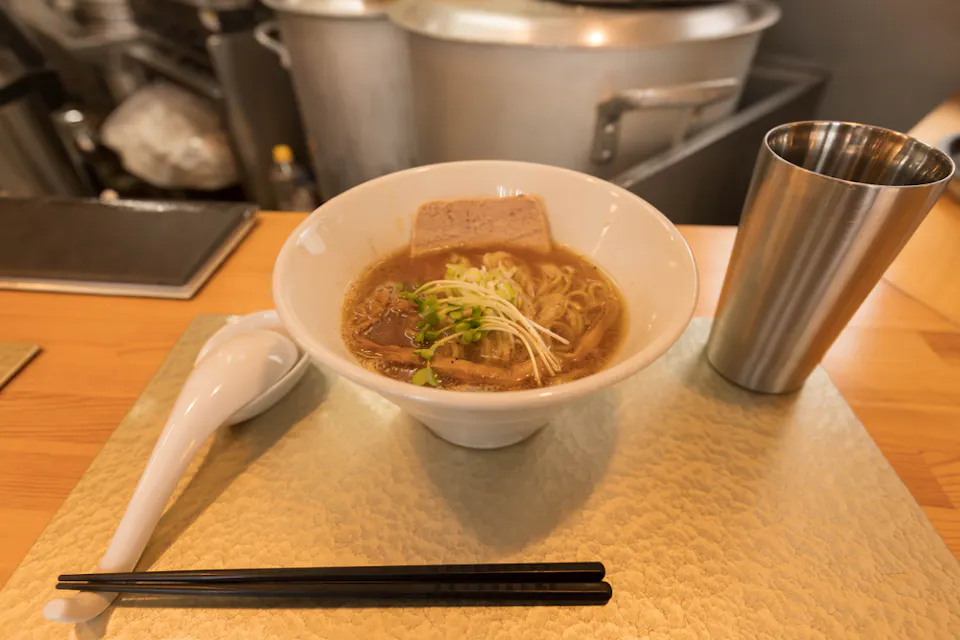 A bowl of ramen placed on a table with a white spoon, black chopsticks, and a stainless steel cup beside it. The ramen features broth, noodles, a slice of meat, bean sprouts, and green onions, with a kitchen background.