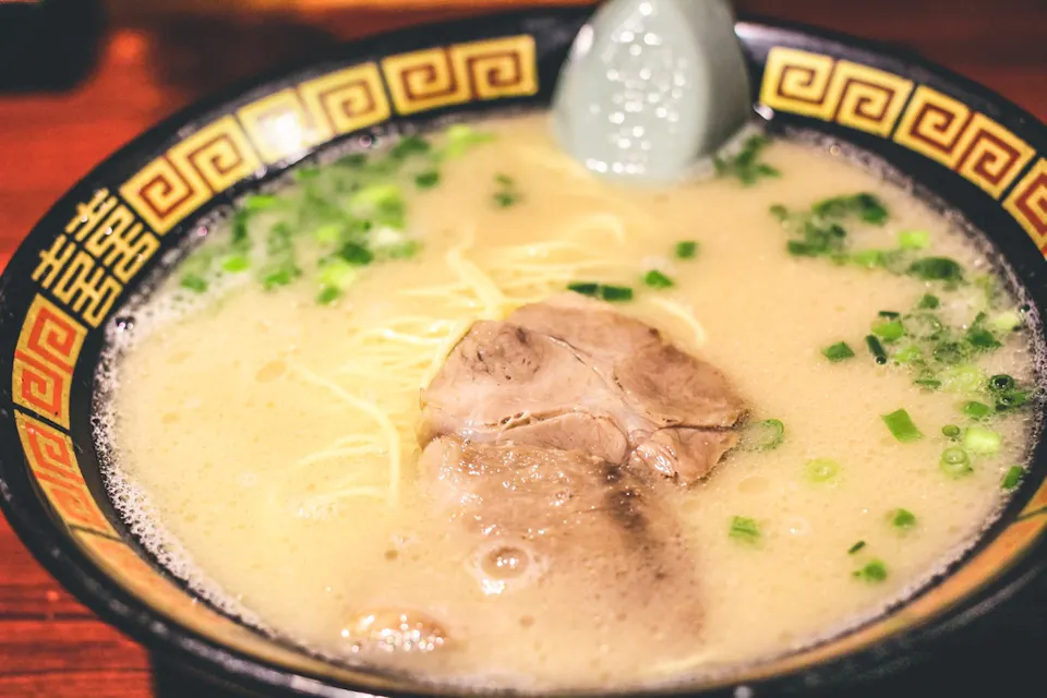 A bowl of ramen soup filled with creamy broth, thin noodles, sliced green onions, and a piece of pork. The bowl has a decorative pattern around the rim, and there is a light blue spoon resting in the soup.
