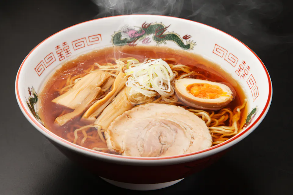 A steaming bowl of ramen with thick broth, topped with slices of pork, half a soft-boiled egg, bamboo shoots, and shredded green onions, served in a bowl decorated with a dragon and Asian-inspired patterns.