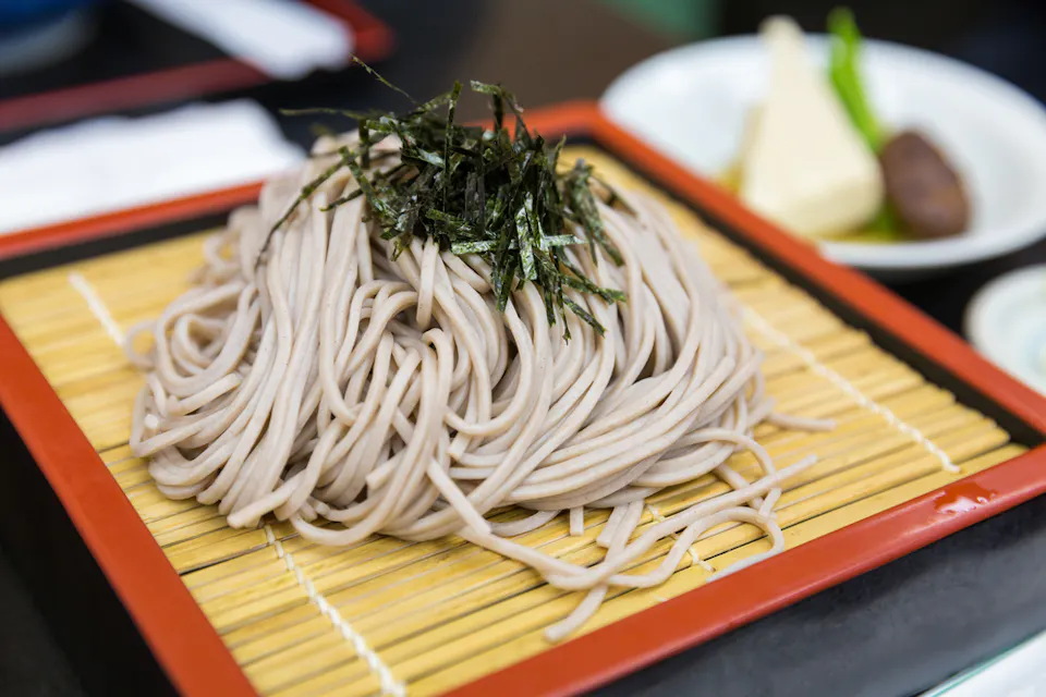 A plate of cold soba noodles topped with shredded nori seaweed. The soba noodles are arranged neatly on a bamboo mat within a red and black rectangular tray. In the background, partially blurred, there is a plate with assorted side dishes.