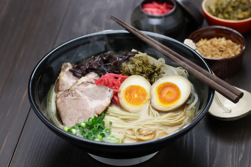 A steaming bowl of ramen is filled with noodles, slices of pork, a halved soft-boiled egg, green onions, pickled ginger, and mushrooms. Chopsticks rest on the bowl's edge. Various side dishes and condiments are blurred in the background.