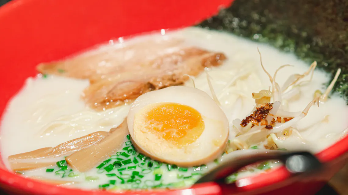 A close-up of a bowl of ramen in a red bowl, featuring a halved boiled egg with a soft yolk, sliced green onions, bamboo shoots, bean sprouts, a slice of pork, and seaweed. The rich broth is creamy, highlighting the various toppings and noodles within.