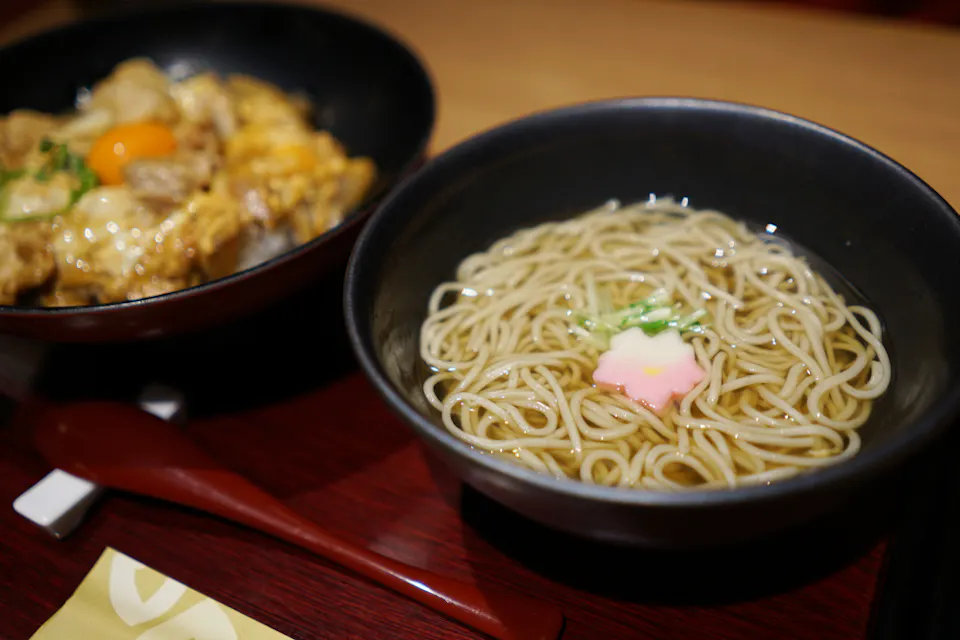 A bowl of ramen noodles in clear broth garnished with a pink and white fish cake and green onions, next to another bowl containing a dish topped with egg and meat, placed on a wooden tray with red chopsticks and a yellow-tipped napkin.