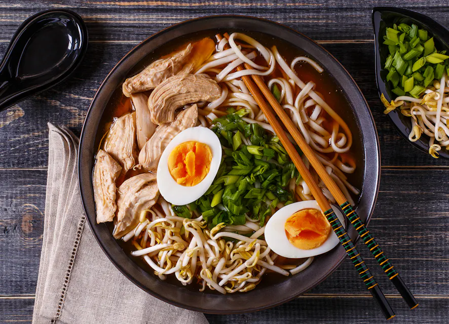 A bowl of ramen with sliced chicken, boiled egg halves, bean sprouts, and chopped green onions, served with wooden chopsticks on a dark wooden table next to a beige napkin and a small bowl of additional green onions.