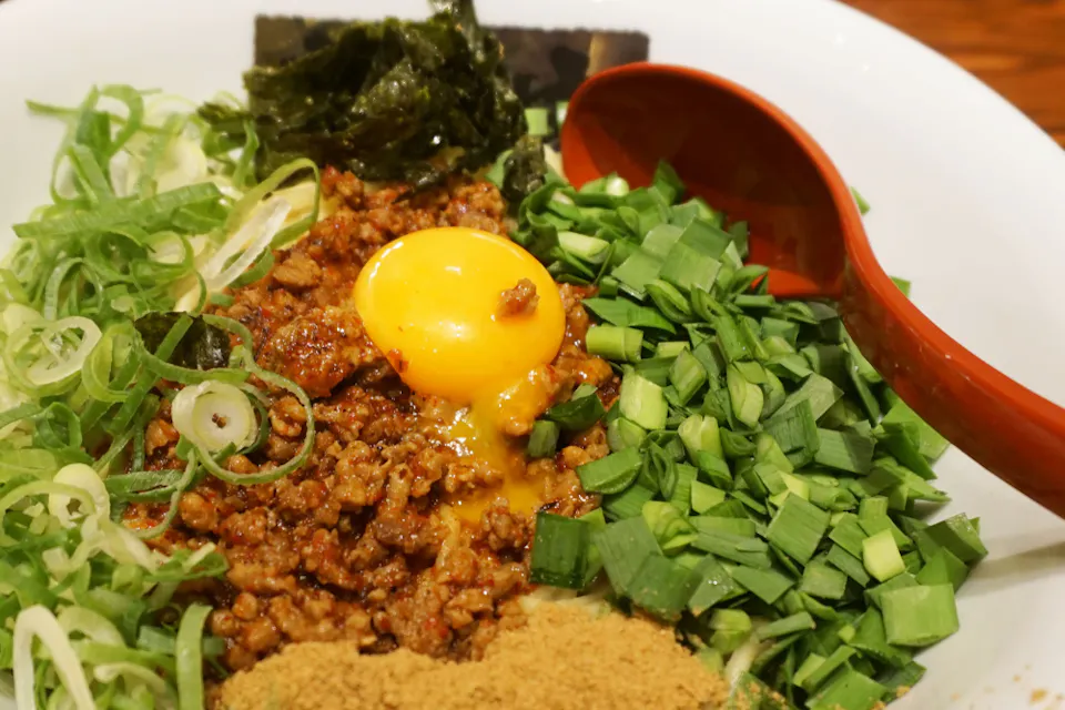 A close-up image of a bowl filled with various ingredients for a savory dish. It includes ground meat, an egg yolk, chopped green onions, fresh greens, and a red spoon resting on the side of the bowl.